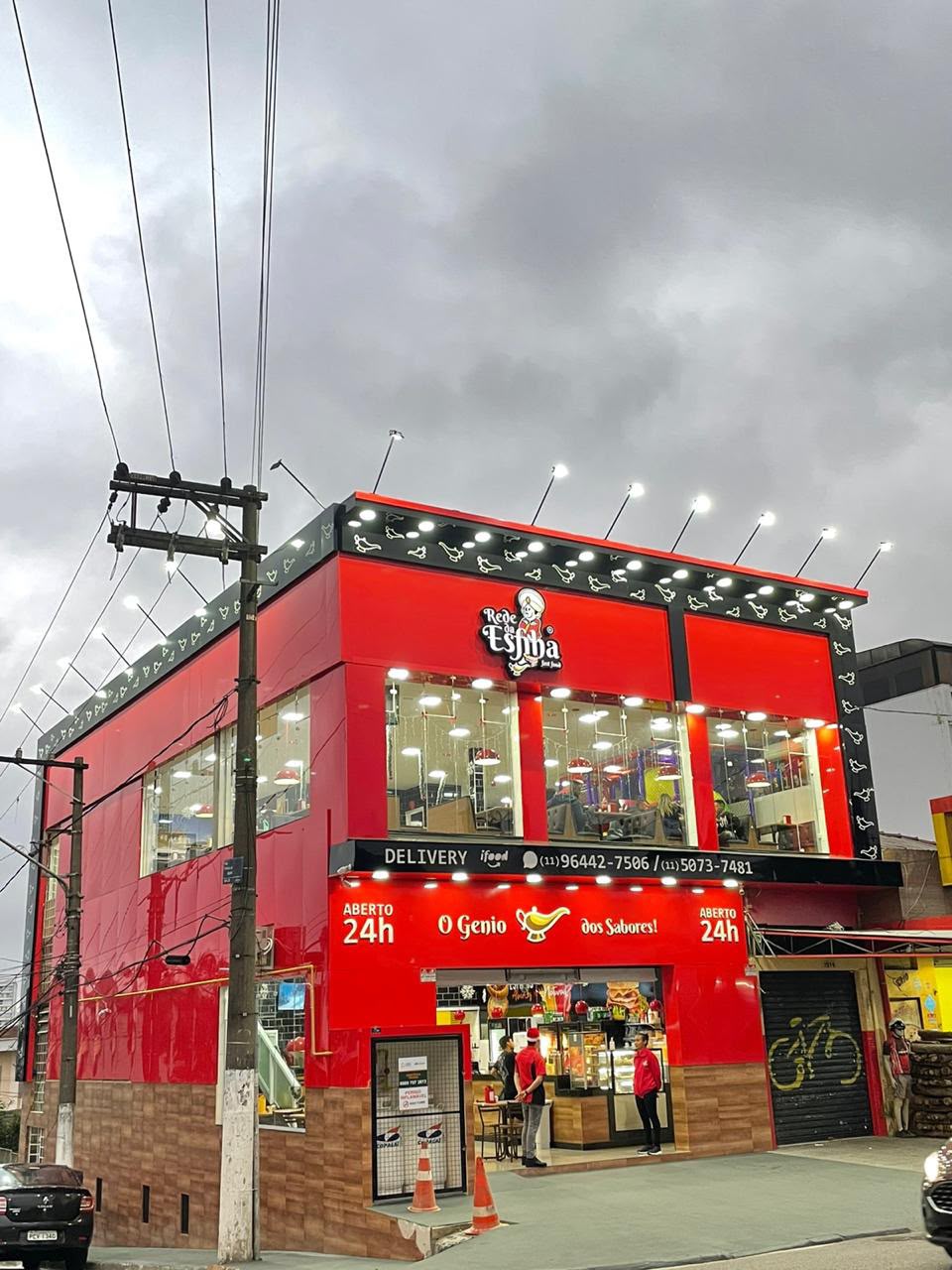Fachada da loja ao anoitecer com placas Aberto 24h e equipe atendendo no balcão